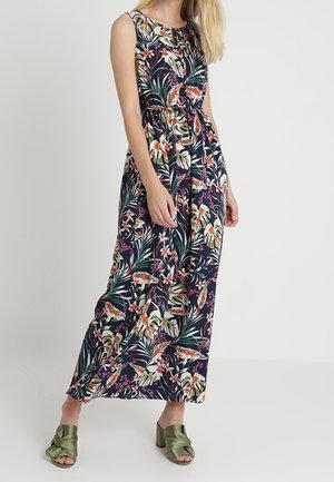 Kvinde iført navyblå langærmet kjole med tropisk blomstermønster og grønne åbne hælede sandaler, stående mod hvid baggrund.