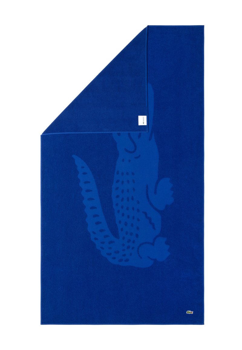 Toalla de algodón azul con un diseño texturizado de cocodrilo en un tono de azul más oscuro, doblada en una esquina, con una pequeña etiqueta de logo.