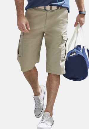Mann trägt beige Cargoshorts, weiße Turnschuhe und ein blaues Hemd, hält eine dunkelblaue Reisetasche mit weißen Trägern.