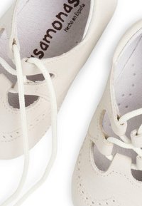 Scarpe in pelle color beige chiaro con ritagli decorativi, suole testurizzate e lacci bianchi, con un'etichetta stampata all'interno che reca la dicitura "Hecho en España."