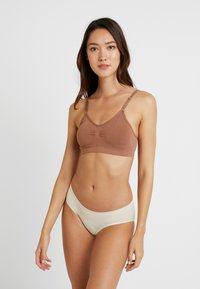Brun bralette med justerbare stropper og subtil samling i midten, parret med kremfargede sømløse truser. Glatt tekstur, enkelt design.