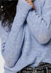 Maglione azzurro in maglia con finitura strutturata e polsini a coste che coprono parzialmente le mani, abbinato a jeans neri e bianchi con motivi a fantasia.
