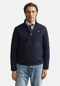 Mornarska prešita jakna z ovratnikom, z osrednjim zadrgo in majhnim logotipom na prsih. Oblečena preko bele srajce in črtaste kravate.