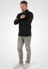 Schwarzer Strickpullover mit hohem Kragen und strukturiertem Muster, kombiniert mit hellgrauen Slim-Fit-Jeans und weißen Sneakers.