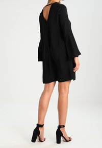 Svart klänning med klockärmar och djup V-back; tillverkad av ett glatt tyg. Matchas med svarta sandaler med blockklack och ankelremmar.