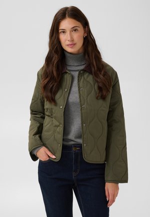 Femme aux longs cheveux bruns portant une veste matelassée vert olive sur un col roulé gris et un jean bleu foncé, debout devant un fond uni.