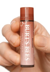 Burt's Bees värviliipar "Red Dahlia," silindrikujuline toru, pruun kork, roosa keha valgete tähtedega, sile tekstuur, hoitakse käes.