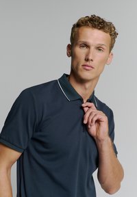Donkerblauwe polo met een ritskraag, voorzien van een contrasterende witte rand. Glad materiaal, korte mouwen en een slank model.