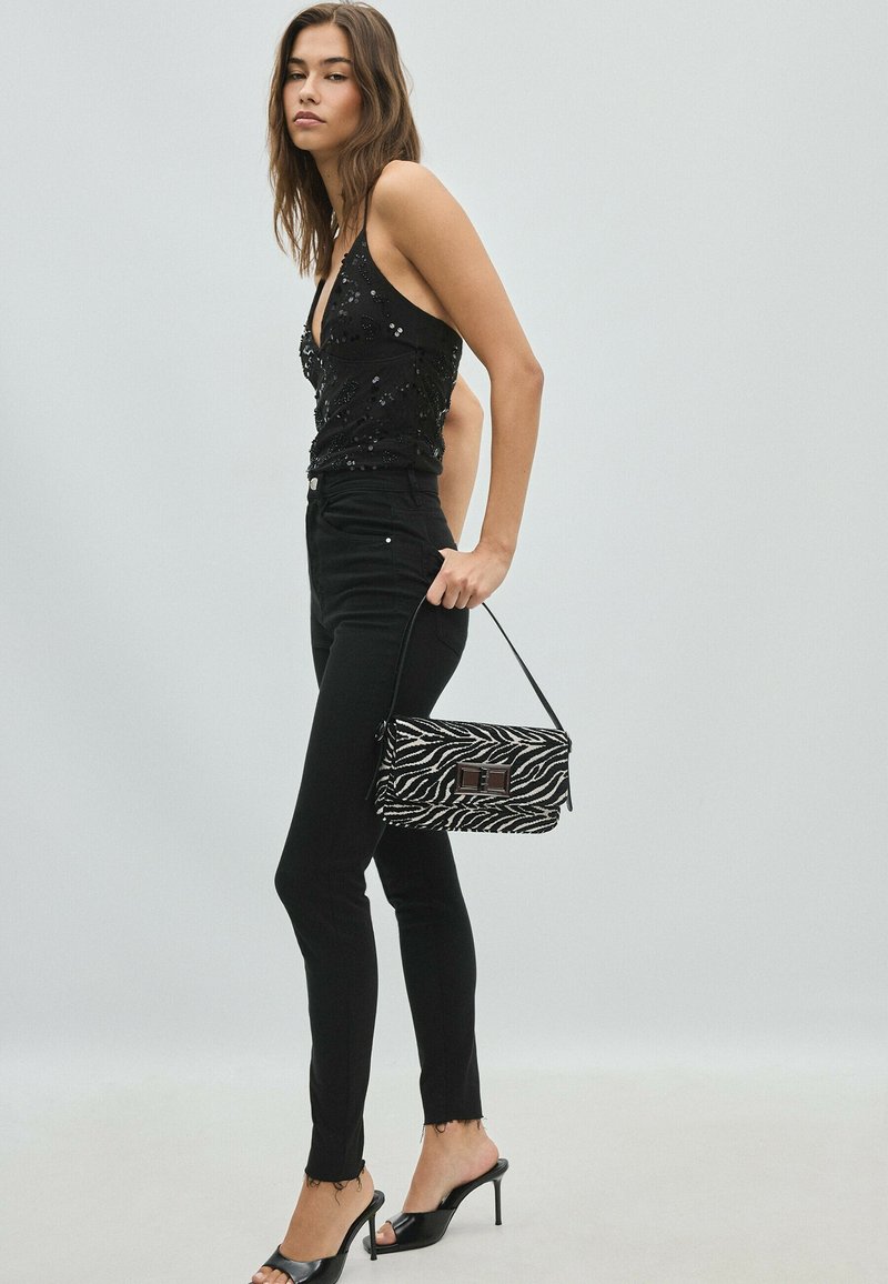 Femme portant un haut noir à sequins, un jean skinny noir et des talons noirs, tenant un petit sac à main imprimé zèbre sur un fond uni.