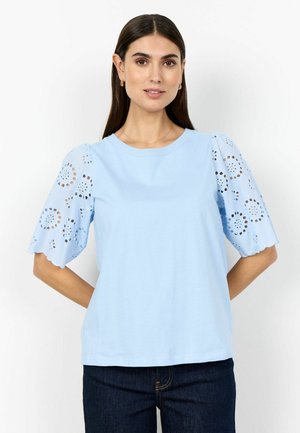 Kvinde iført lyseblå top med hulmønstrede pufærmer og mørke jeans, stående mod en ensfarvet hvid baggrund