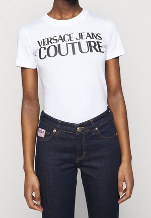 Personne portant un t-shirt blanc avec le texte « VERSACE JEANS COUTURE » et un jean taille haute bleu foncé avec une étiquette rose sur la poche.