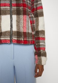 Giacca in pile con zip caratterizzata da un motivo a quadri rosso, marrone e bianco, con una finitura morbida e texturizzata. Abbinata a pantaloni blu chiaro.