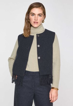 Frau mit schulterlangem, hellbraunem Haar, die goldene Creolen trägt, einen beigen, gerippten Rollkragenpullover, eine marineblaue, knöpfbare Weste mit Fransen und marineblaue Hosen.