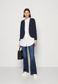 Blazer blu navy con due bottoni sopra una camicia bianca, abbinato a jeans svasati in denim e sneakers bianche; tiene in mano una borsa nera.