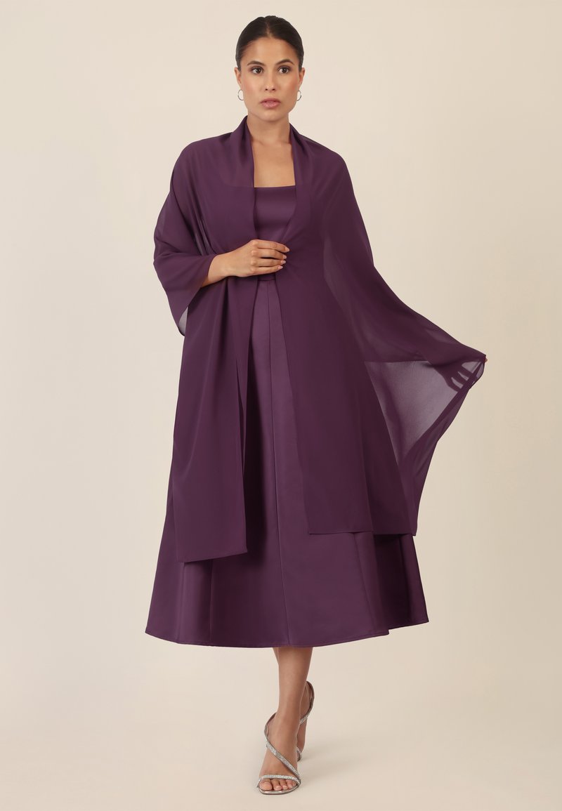 Lila schichtige Kleid mit einem passenden Schal; seidige Textur, ärmelloses Design und ein fließender Rock, der knapp unter dem Knie endet. Ergänzt durch silberne Sandalen.