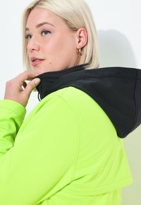 Nainen, jolla on lyhyet vaaleat hiukset, säätää neonvihreän takin kaulusta, jossa on musta huppu, tasaisen valkoista taustaa vasten.