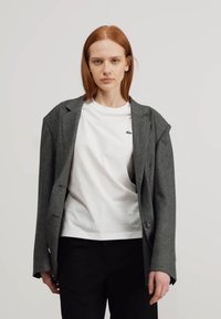 Grau gestreifter Blazer mit Reverskragen, Ein-Knopf-Verschluss und Schulterpolstern, getragen über einem weißen T-Shirt und schwarzen Hosen.