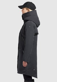 Black, long winter coat with a high collar, zippered pockets, and side vents; features a matte finish and embossed logo on sleeve.
