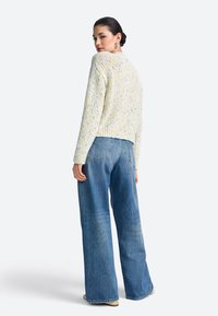 Übergroßer cremefarbener Pullover mit bunten Farbsprenkeln, kombiniert mit hoch taillierter blauer Jeans, die eine legere Passform und gerippte Bündchen zeigt.