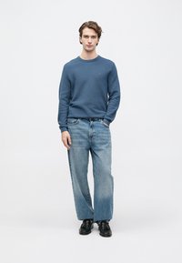 Jauns vīrietis stāv ar zilu garām piedurknēm džemperi, gaiši ziliem platām kājām džinsiem un melnām ādas kurpēm uz vienkārša balta fona.