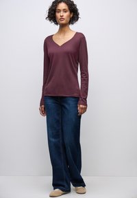 Burgunderfarbene Bluse mit langen Ärmeln und V-Ausschnitt, kombiniert mit weit geschnittenen, dunkelblauen Jeans und beigen Schuhen. Weicher Stoff mit lockerer Passform.