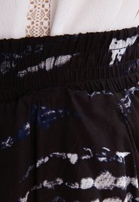 Tissu noir avec un motif éclaboussé en blanc et bleu. Présente une taille élastique et une texture douce. Vue en gros plan des détails du vêtement.