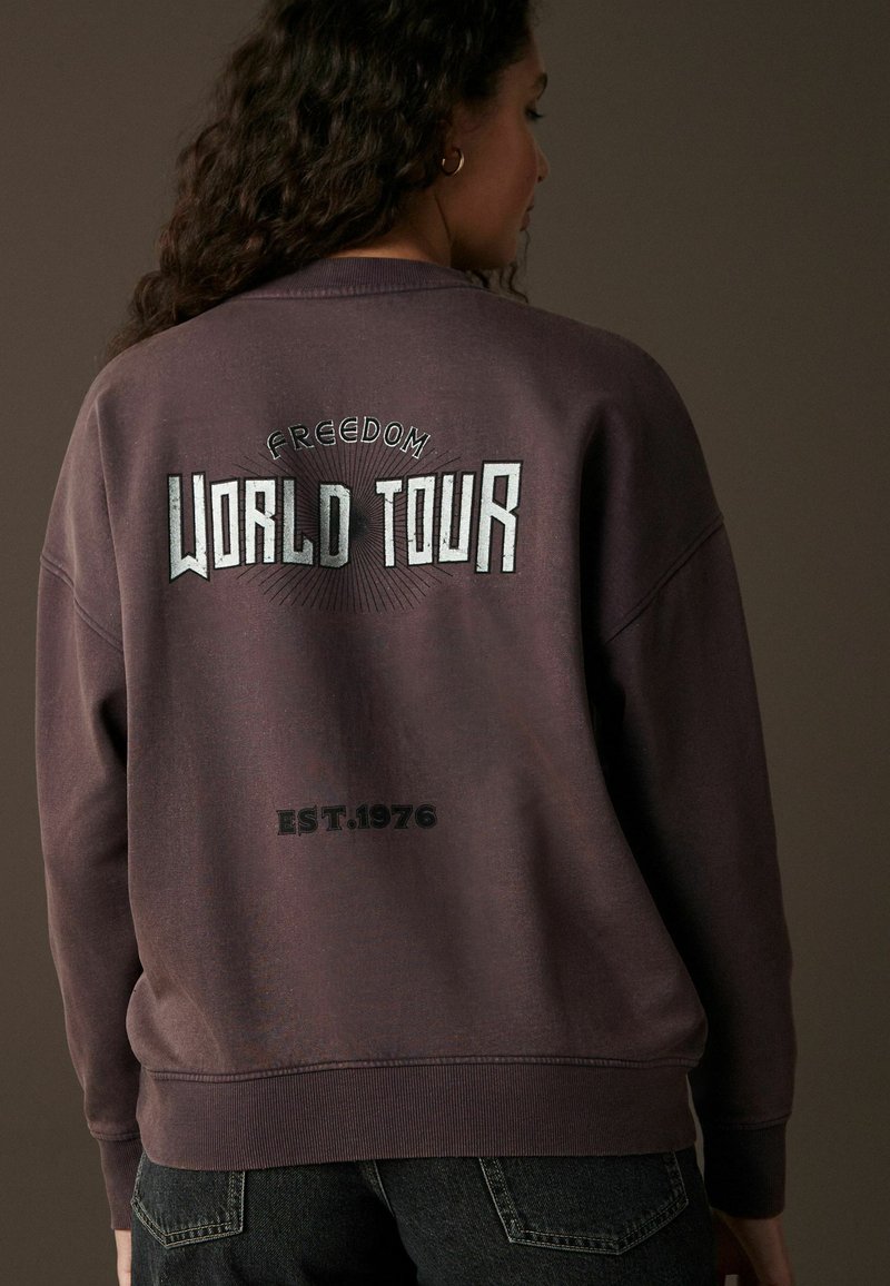 Sweatshirt violet avec "FREEDOM WORLD TOUR" en lettres blanches dans le dos, avec "EST. 1976" en dessous ; tissu doux et coupe ample.