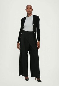 Cardigan nero con bottoni dorati, indossato sopra una maglietta grigia chiara. Pantaloni larghi neri e scarpe con tacco completano l'outfit.