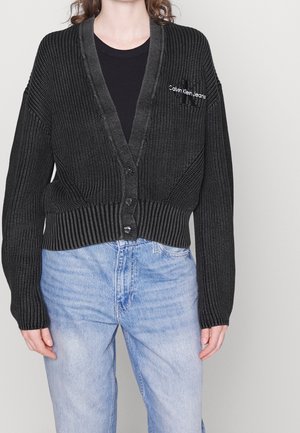 Osoba ubrana w czarny dzianinowy kardigan na guziki, z logo Calvin Klein Jeans na piersi, nałożony na czarną koszulę, w połączeniu z jasnoniebieskimi dżinsami.