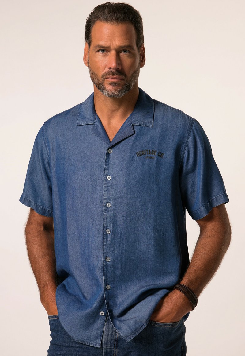 Homme aux cheveux courts et foncés et à la barbe, portant une chemise bleue à manches courtes avec le texte "Heritage Co", mains dans les poches, fond neutre.