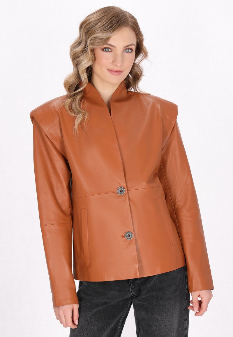 Braune Lederjacke mit hohem Kragen, oversized Schultern, zwei seitlichen Taschen und einem einzigen Knopfverschluss. Kombiniert mit schwarzen Jeans.