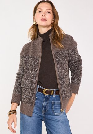 Woman wearing brown patterned zip-up cardigan, dark turtleneck, blue jeans, gold hoop earrings, and bracelets against a plain background.