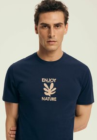 Granatowa koszulka z krótkim rękawem wykonana z bawełny, z jasną grafiką na środku, na której znajduje się napis "CIESZ SIĘ PRZYRODĄ" oraz motyw liścia.