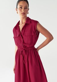 Abito rosso senza maniche con bottoni sul davanti, due tasche sul petto e vita regolabile con coulisse, realizzato in tessuto morbido.