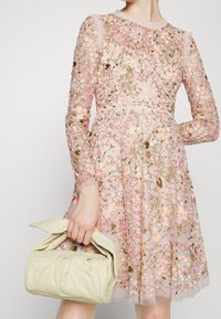 Robe rose clair brodée de motifs floraux, en tissu transparent, avec des manches longues. Sac à main beige plissé tenu à la main.