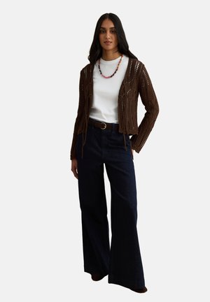 Donna che indossa un cardigan marrone a maglia aperta, t-shirt bianca, pantaloni scuri a gamba larga, cintura marrone e collana colorata di perline, in piedi con la mano in tasca.