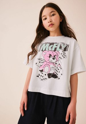 Junges Mädchen mit langen dunklen Haaren, das ein weißes T-Shirt mit einem pinken Bären, Würfeln und dem Schriftzug „MCFLY“ trägt, kombiniert mit dunkler Hose.
