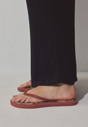 Stopy w czerwonych japonkach z wzorzystymi podeszwami, stojące na jasnej podłodze, czarne spodnie sięgające kostek.