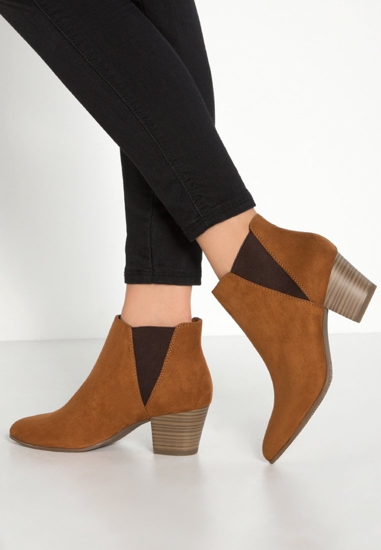 Femme portant un pantalon noir ajusté et des bottines en daim marron avec talons empilés et panneaux latéraux élastiques.