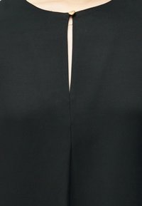 Μαύρη μπλούζα με στρογγυλή λαιμόκοψη, με λεπτομέρεια «κλειδώματος» που κλείνει με ένα χρυσό κουμπί, κατασκευασμένη από λεία ύλη με διακριτική γυαλάδα.