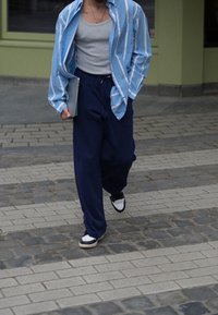 Camisa de botones a rayas azul claro y blanca sobre una camiseta sin mangas gris, combinada con pantalones anchos azul marino y zapatillas negras y blancas. Sosteniendo una computadora portátil.