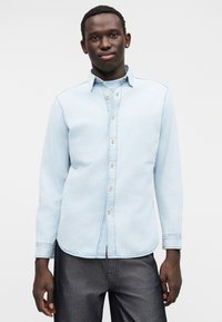 Camisa de ganga azul clara com colarinho de botões, mangas compridas e abotoamento frontal. Apresenta costura contrastante e bainha curva.