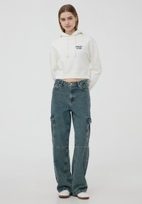 Felpa bianca corta con cappuccio, cordoncini e dettagli del logo, abbinata a jeans cargo blu con tasche laterali e design a gamba larga.