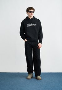 Μαύρο φούτερ με κουκούλα και λογότυπο «Dickies», μαύρο φαρδύ παντελόνι και γκρι παπούτσια. Το μοντέλο φορά γυαλιά ηλίου. Ύφασμα με ομαλή υφή, αέρινη εφαρμογή, μινιμαλιστικός σχεδιασμός.