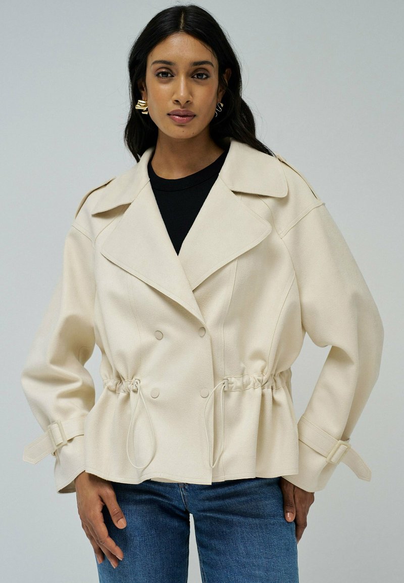 Mujer con una chaqueta crema de doble botonadura, con cintura ajustada y correas con hebillas en las mangas, combinada con vaqueros azules y un top negro.