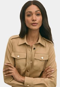 Camicia button-up marrone realizzata in tessuto morbido, caratterizzata da mostrine, due tasche sul petto e bottoni color oro. Maniche lunghe con una vestibilità rilassata.