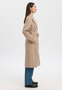 Kvinne i beige belagt ullfrakk, blå jeans og svarte loafers står i profil mot en enkel lys bakgrunn.