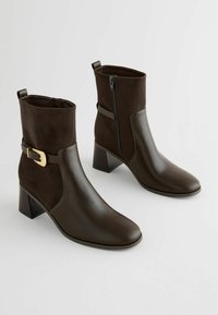 Bottines marron en cuir et daim, dotées d'une fermeture éclair, d'une boucle décorative et d'un talon carré. Texture lisse avec une finition mate.