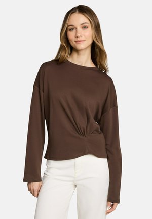 Jeune femme aux cheveux châtain clair porte un haut marron à manches longues avec un détail froncé et un pantalon blanc sur un fond uni.