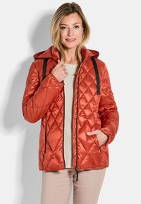 Gequilted, gewatteerde oranje jas met een capuchon; heeft zwarte ritsen en banden, en een gestructureerde, glanzende afwerking. Draagt over een crèmekleurige trui.